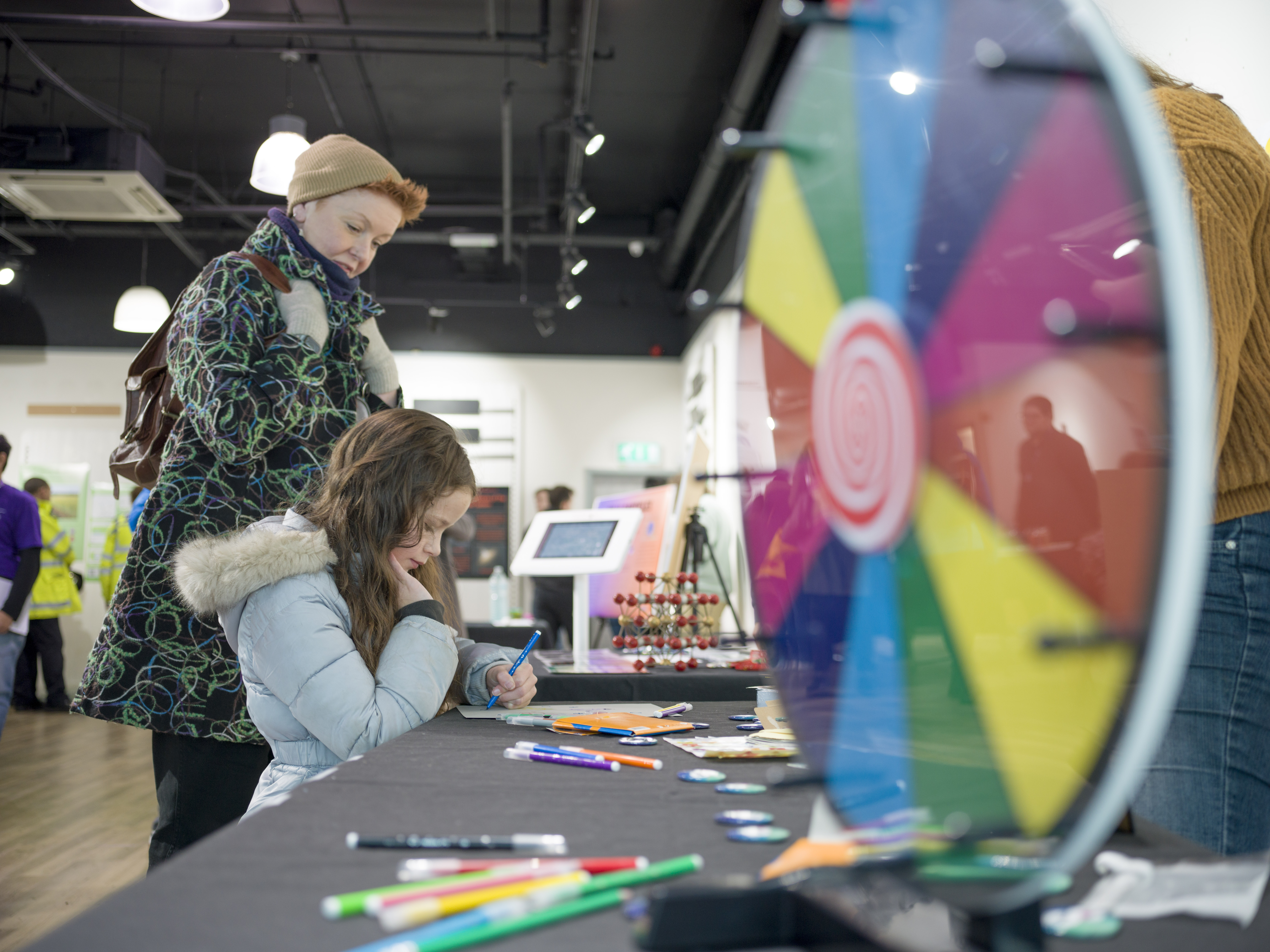 A child drawing with a coloured pen. There is a multi-coloured spinner on the table. An adult is watching. 