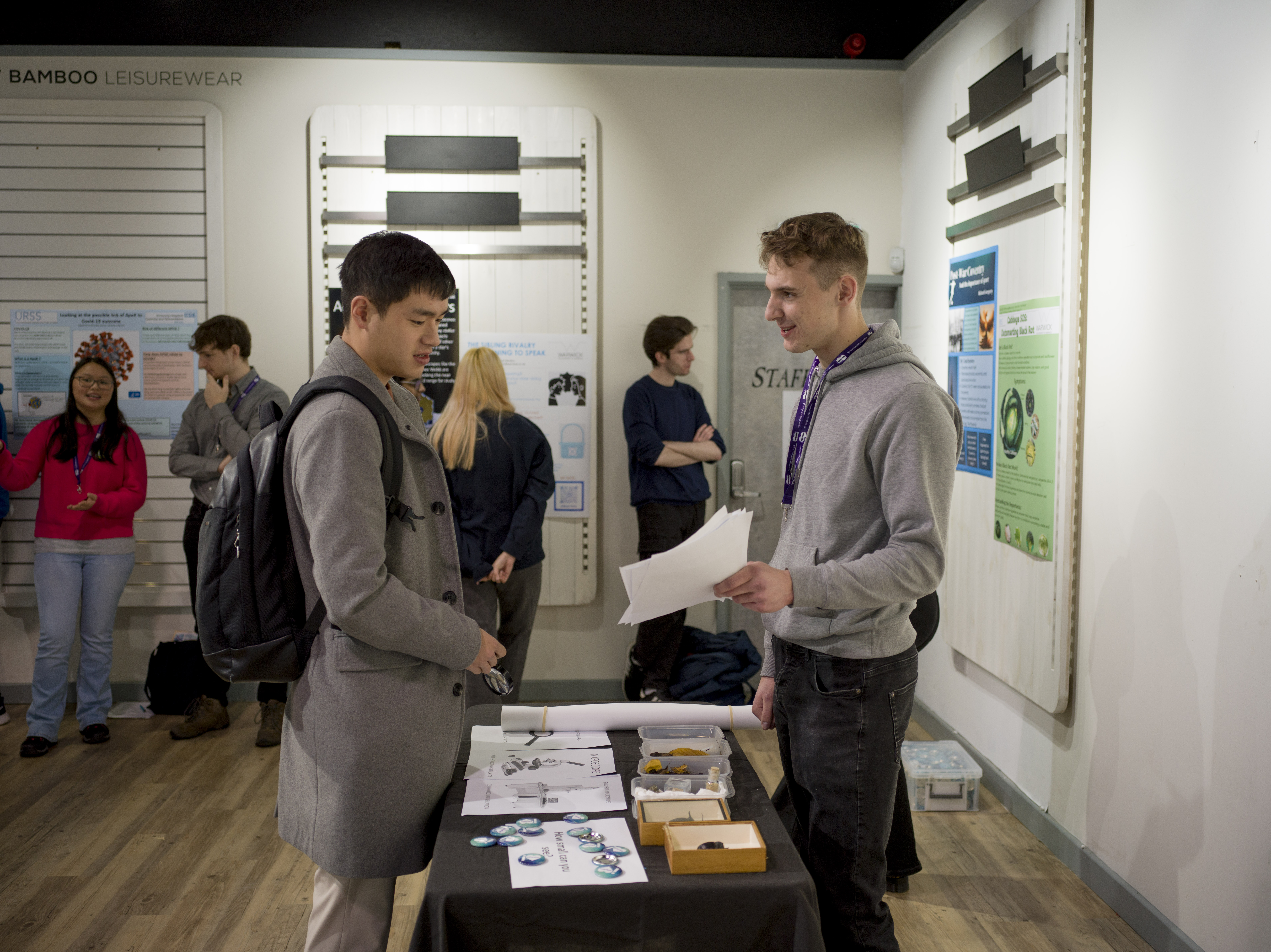A student shows pictures to a visitor. On the table are various objects, including large models of insects in boxes. 