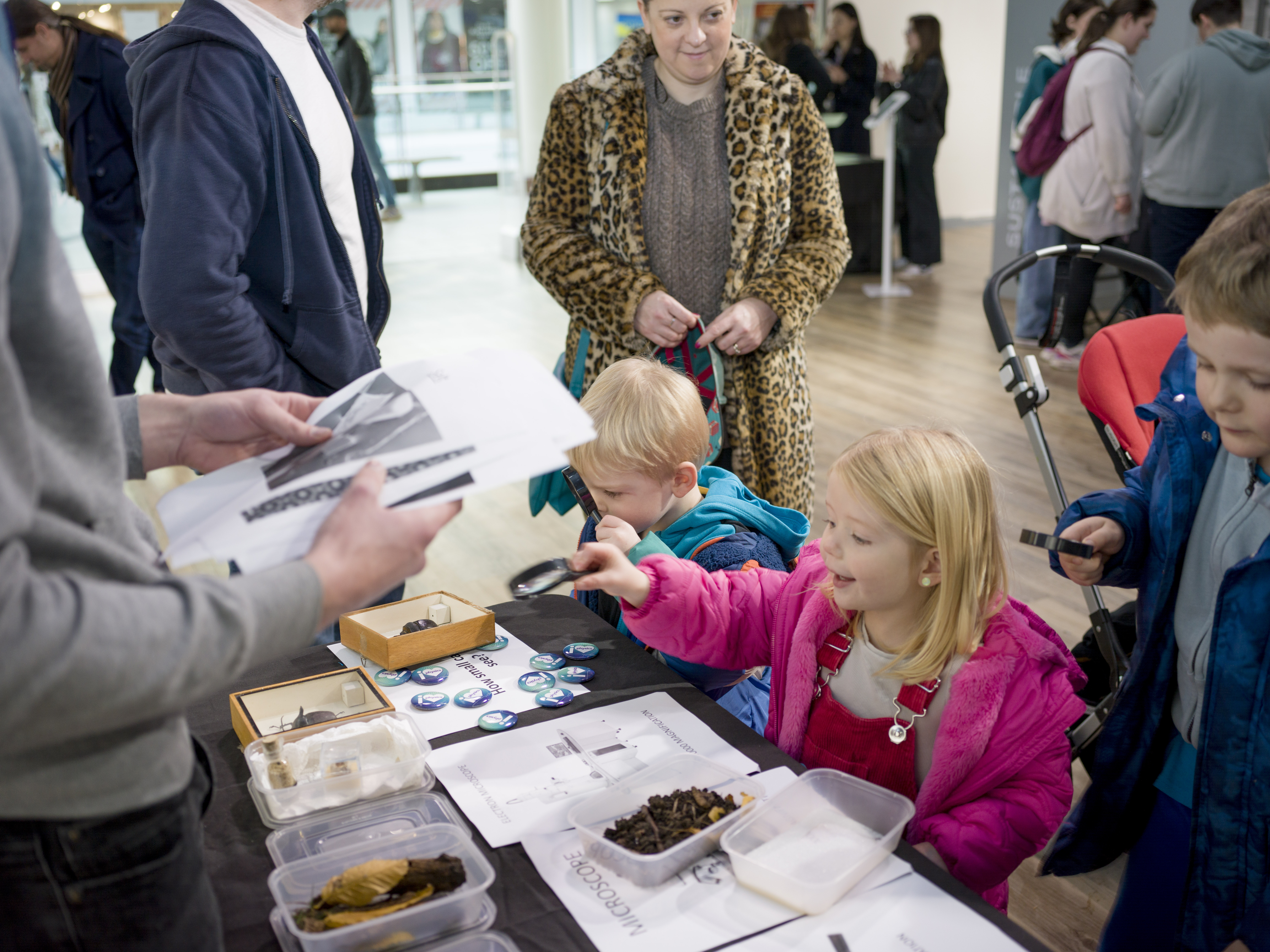 Three children using microscopes to look at different objects on a table. Some adults are watching them. 