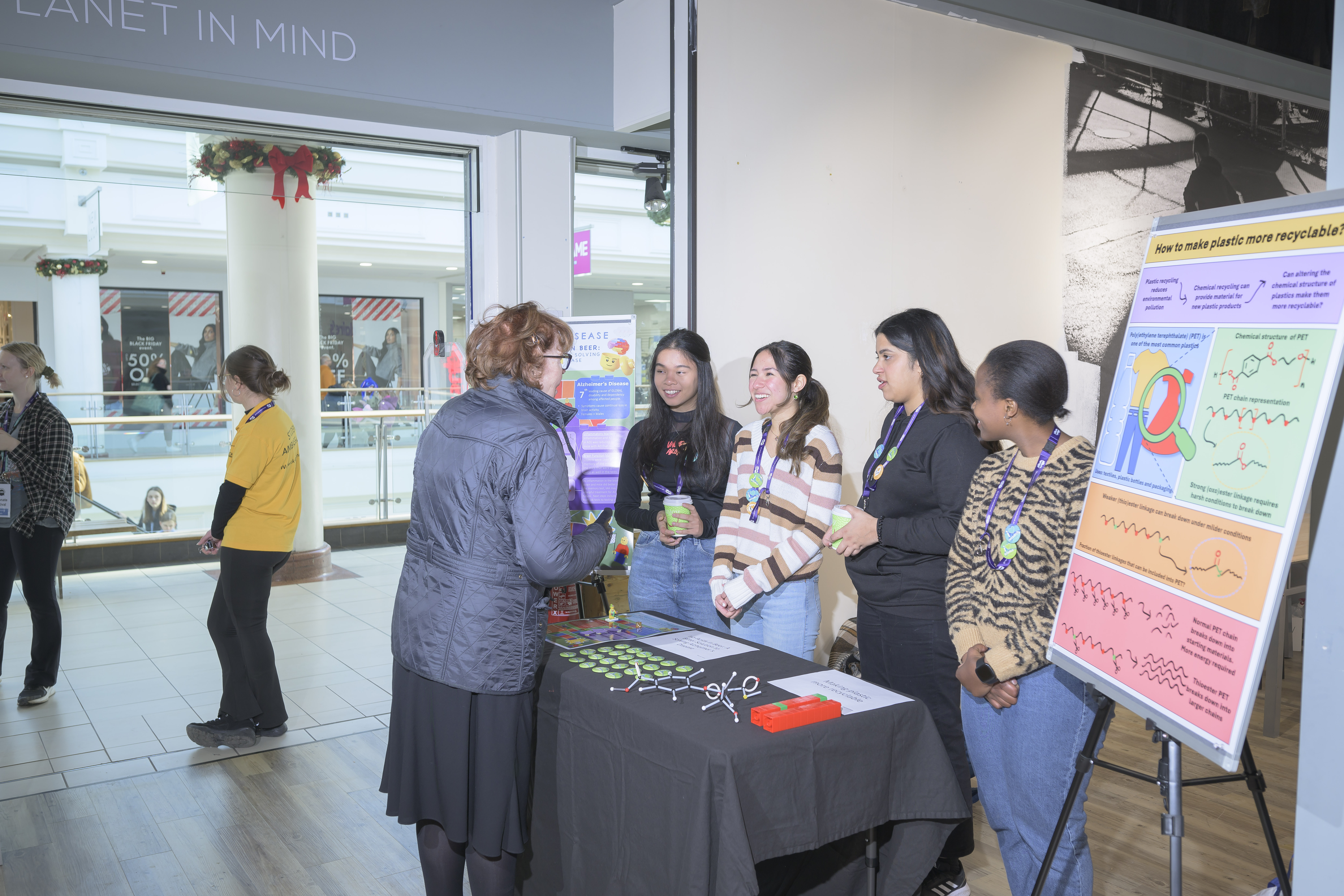 Four students smile as they listen to a visitor. They are standing between two posters and a table. 