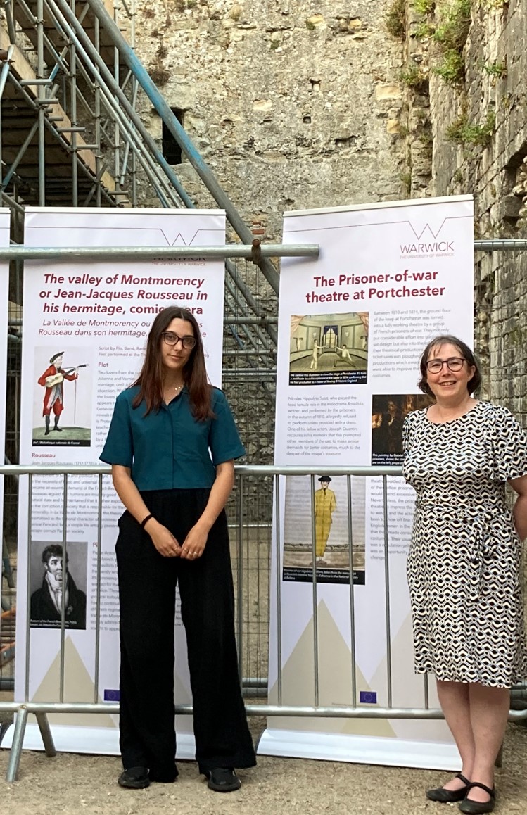 Eve and Kate in the castle, standing in front of two tall, white banners with text and images on them. 
