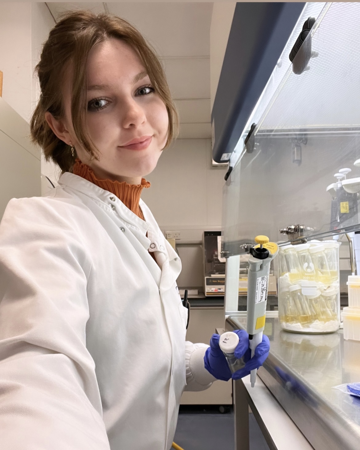 A photo of Arianwen in a lab. She's wearing a white lab coat and blue gloves.