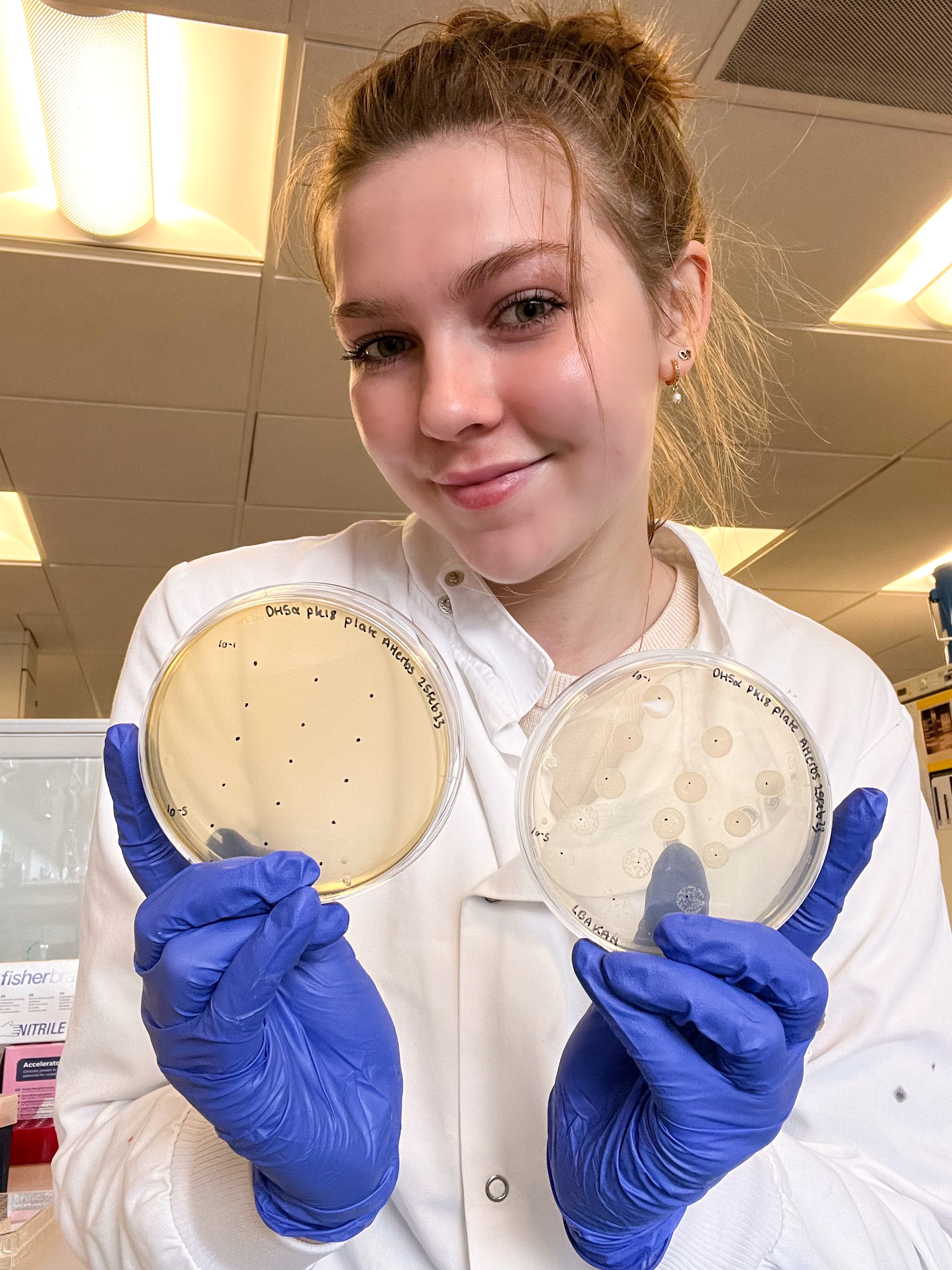 Arianwen holding two petri dishes. She is wearing a white lab coat and blue gloves.