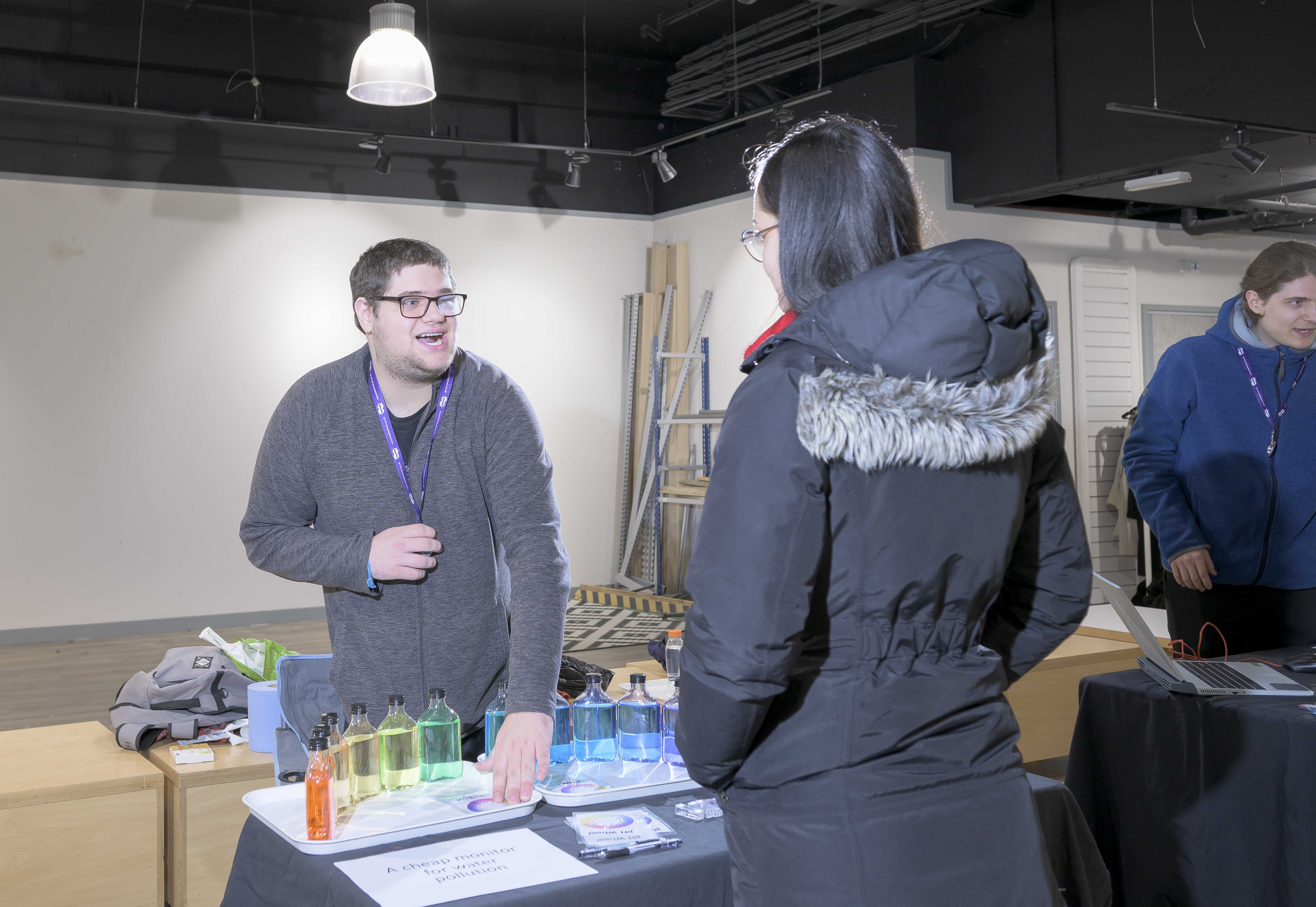 A student talking to a visitor. On the table between them are bottles of different coloured liquids, from red to blue. 