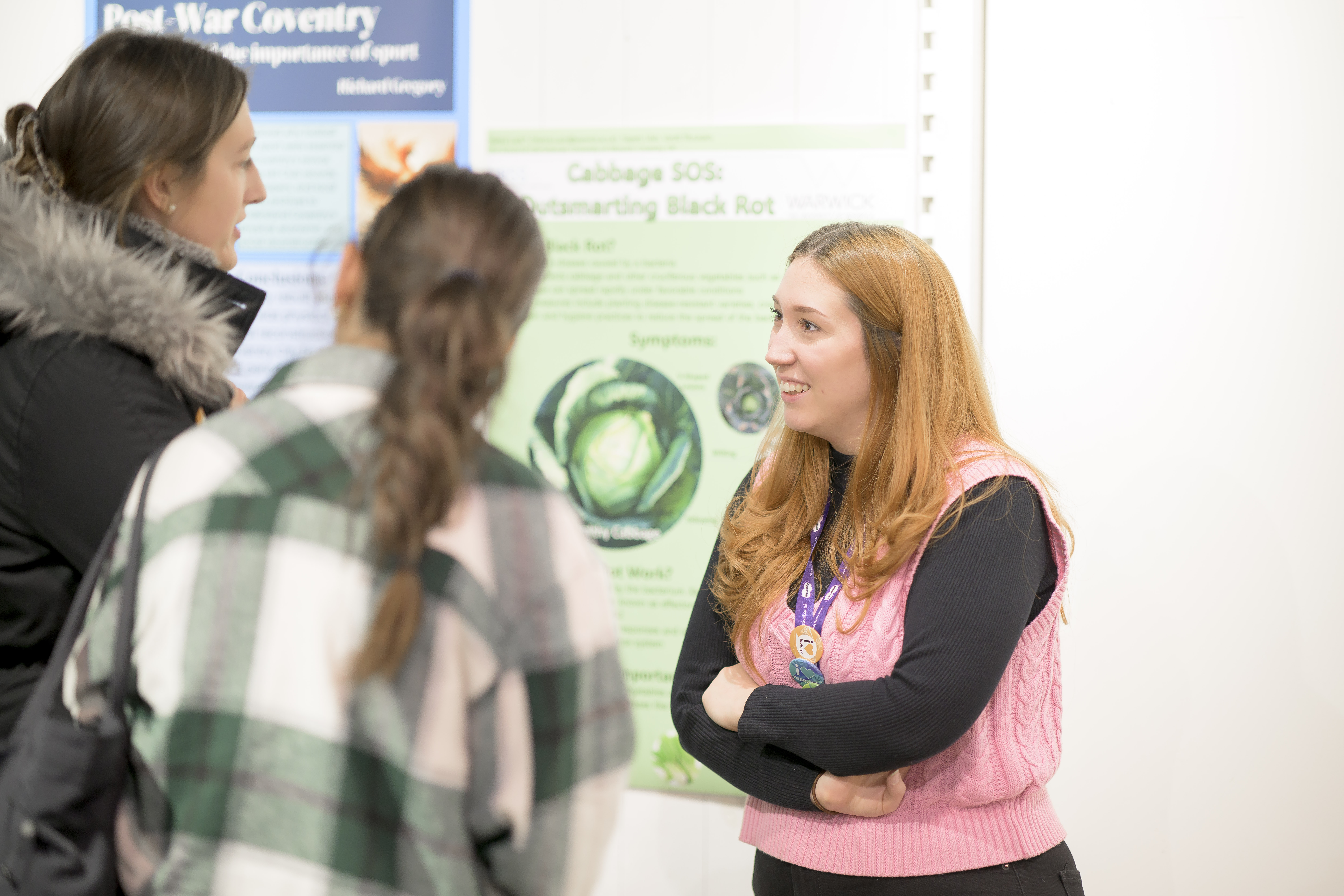 A student smiling as they listen to two visitors in front of a green poster. 