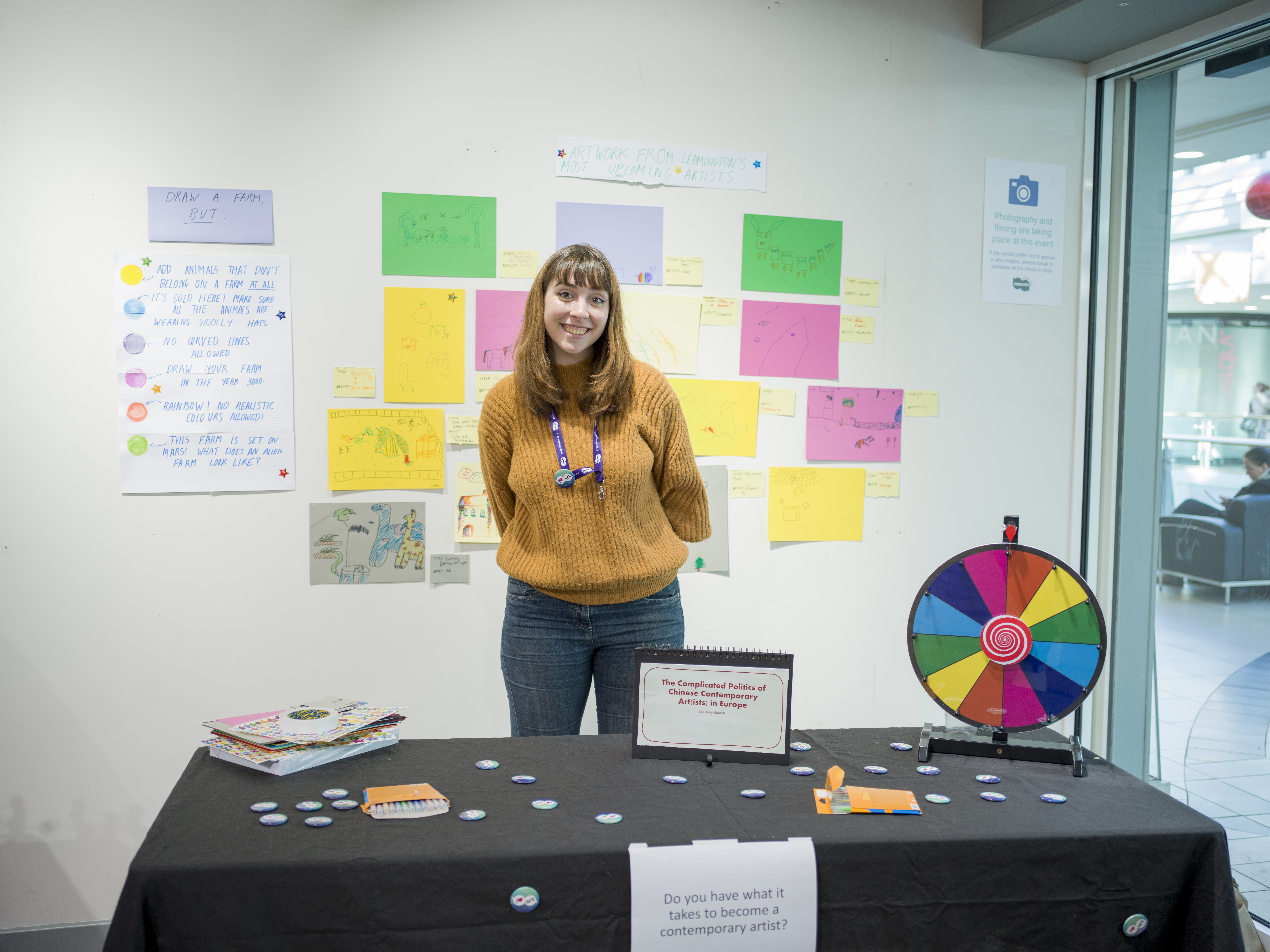 A student standing behind a table with coloured paper and pens and a multi-coloured spinning wheel. On the wall in the background are lots of pictures. 