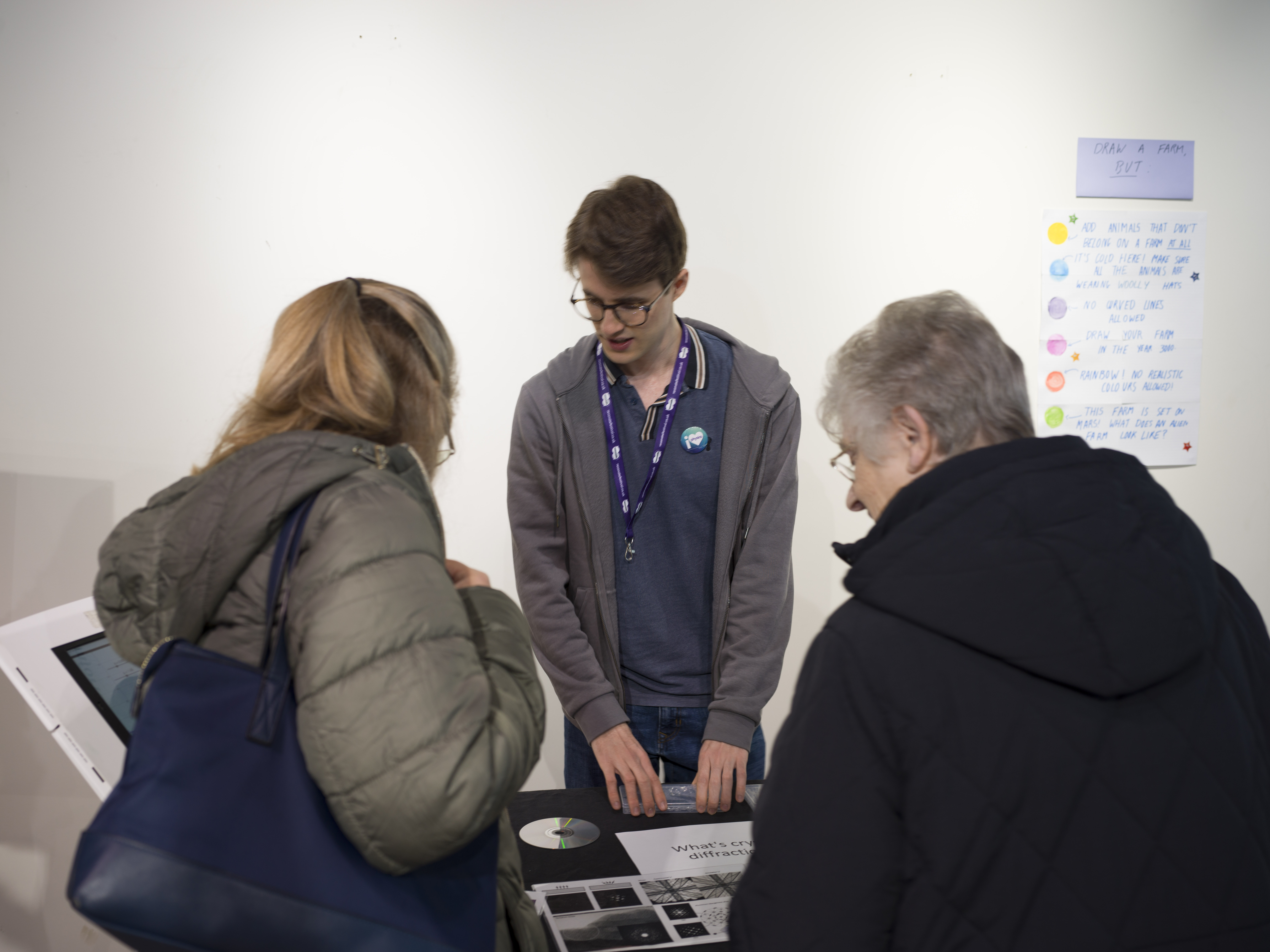 A student talking to two members of the public. They have a table with objects on it, including a CD, and an iPad on a stand. 
