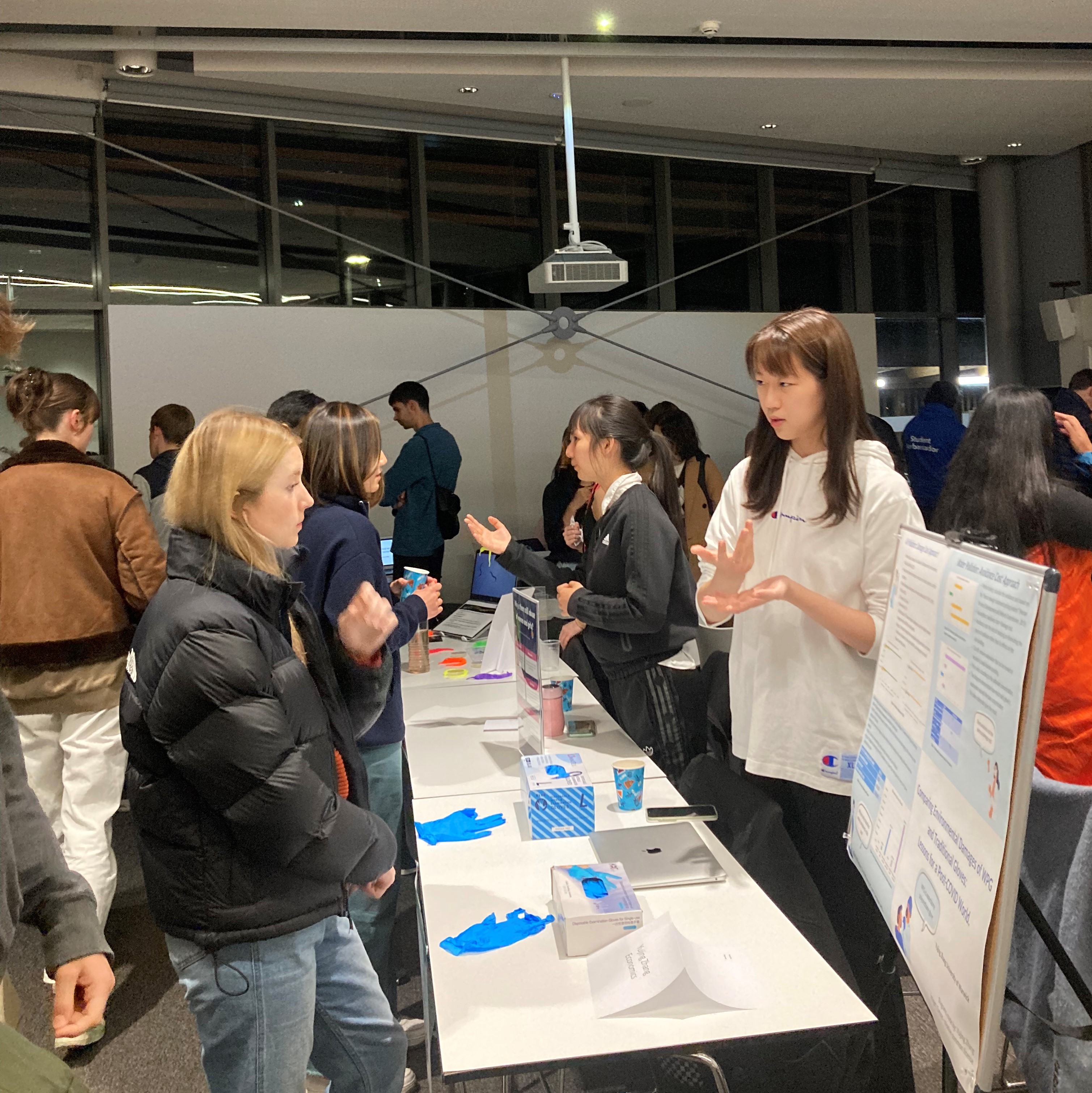 In the foreground, two students are standing on either side of a table and talking to each other. On the table are two kinds of blue gloves. In the background are more students and visitors talking over tables.