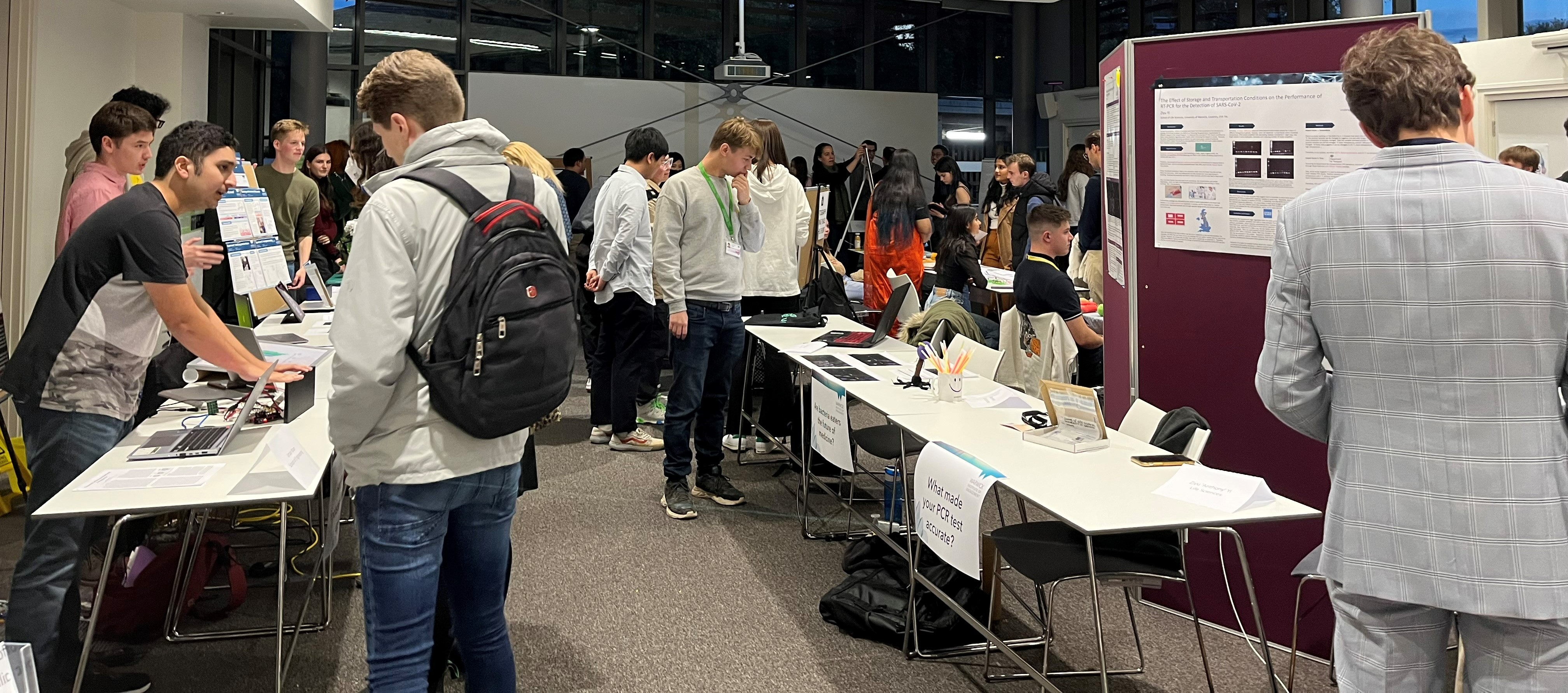 Tables set out in a market style with students standing behind them - each student is talking about a different project with a visitor looking at their poster or physical resources.