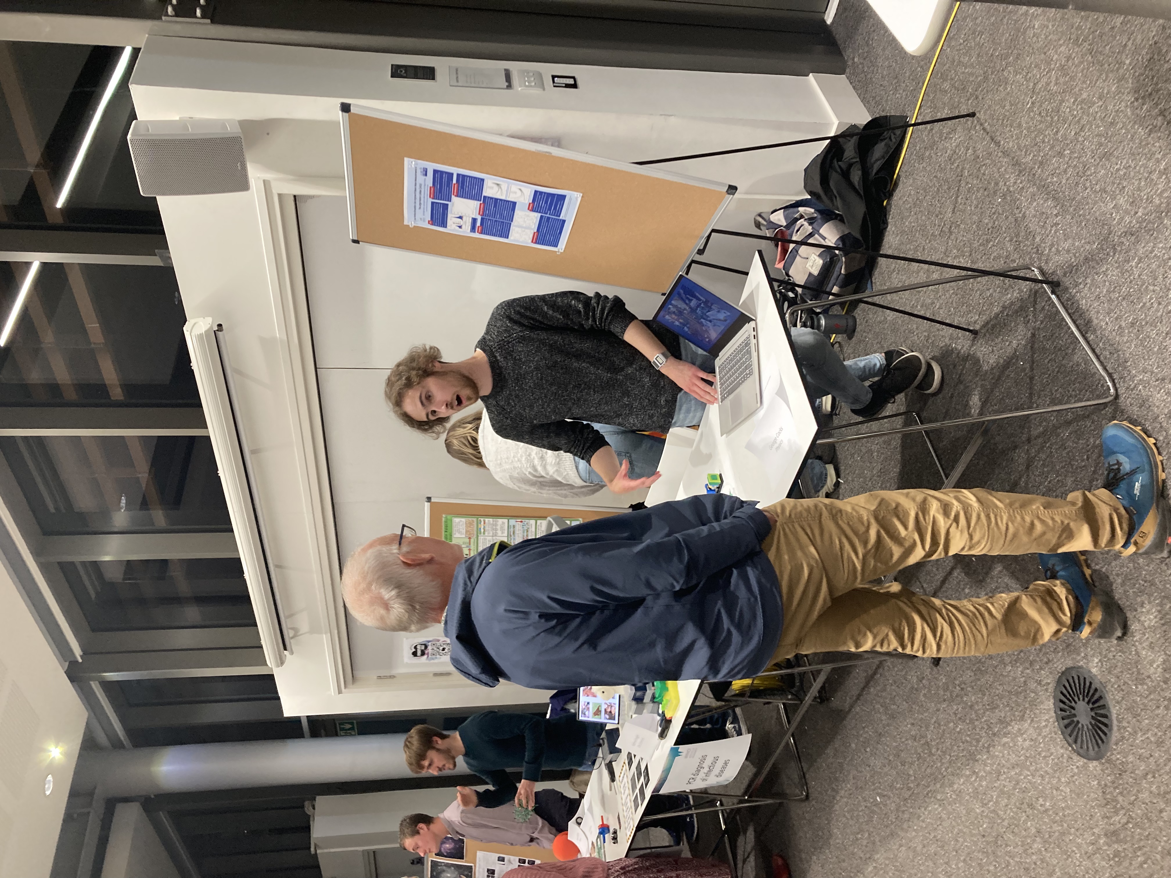 A student stands behind a table, with a poster next to them on a board and a laptop screen display on the table. They are talking to a visitor. In the background, other students are doing the same at their tables. 