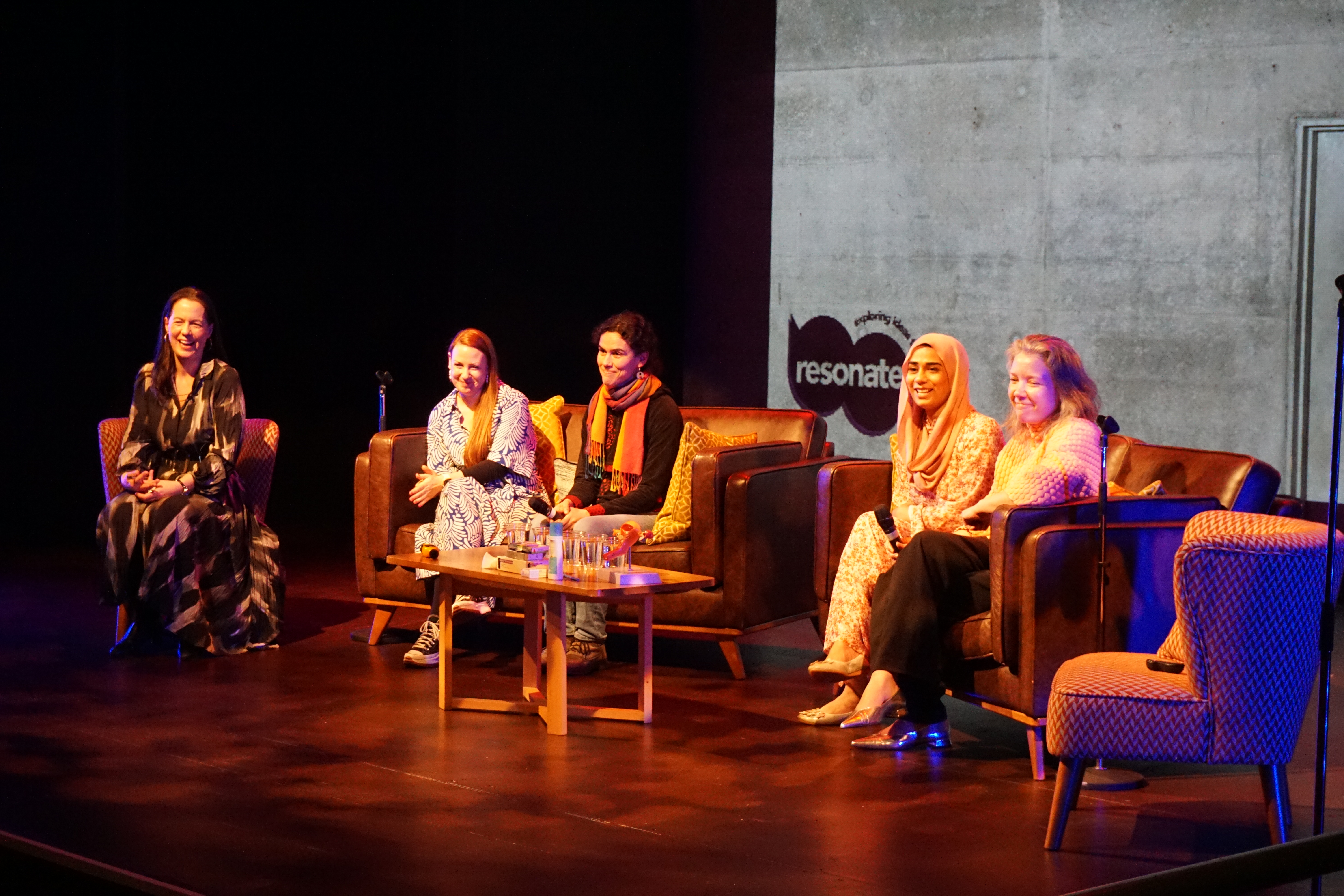 Five smiling people sitting on sofas and an armchair on a stage. 